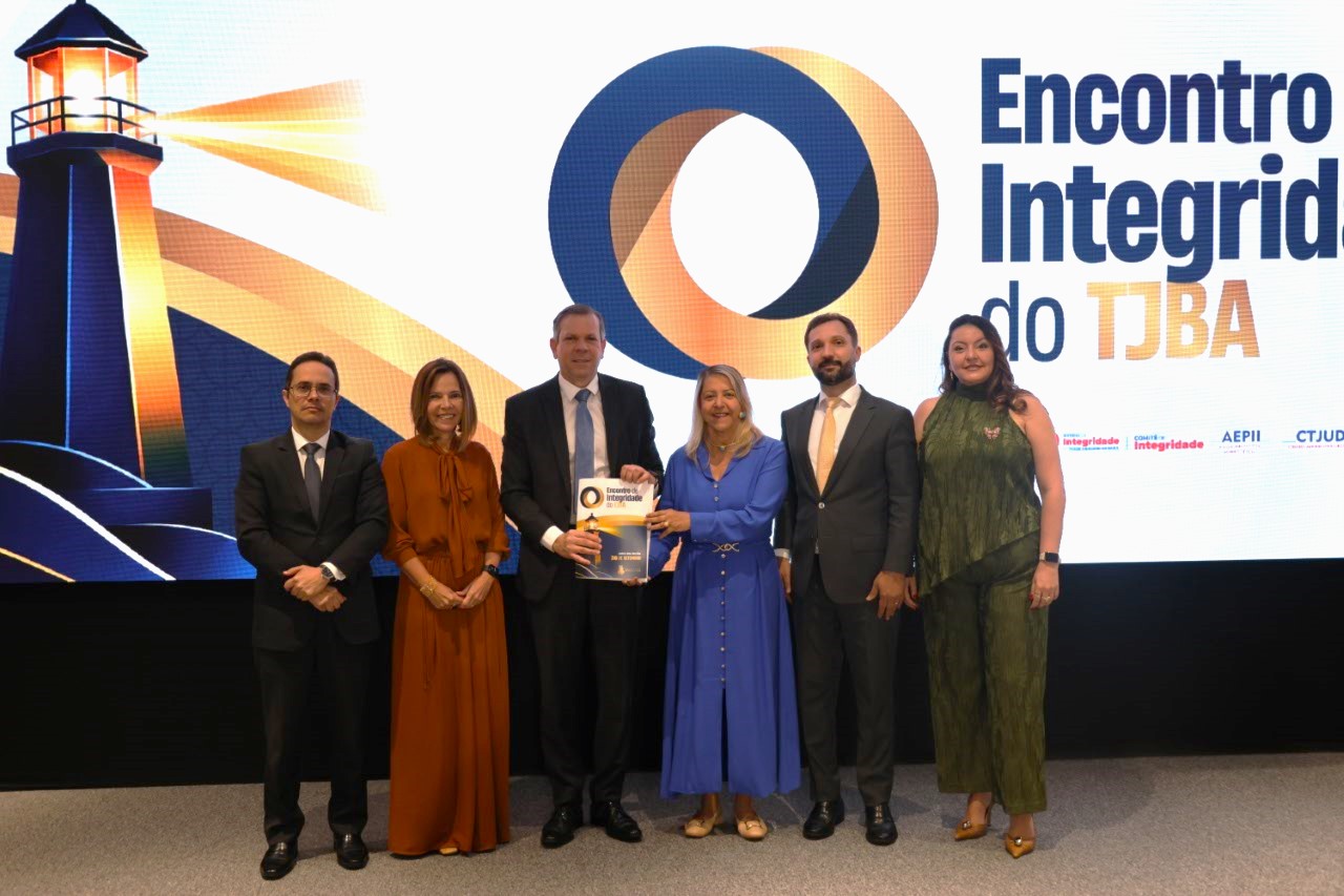 Seis pessoas estão em pé, lado a lado, diante de um grande painel iluminado. Ao fundo, aparece a imagem de um farol, um símbolo circular em azul e laranja e o texto “Encontro de Integridade do TJBA”. As pessoas usam roupas sociais; duas vestem ternos, três usam vestidos e uma usa blusa e calça. A pessoa ao centro segura um certificado ou material do evento. Todos posam para a foto.