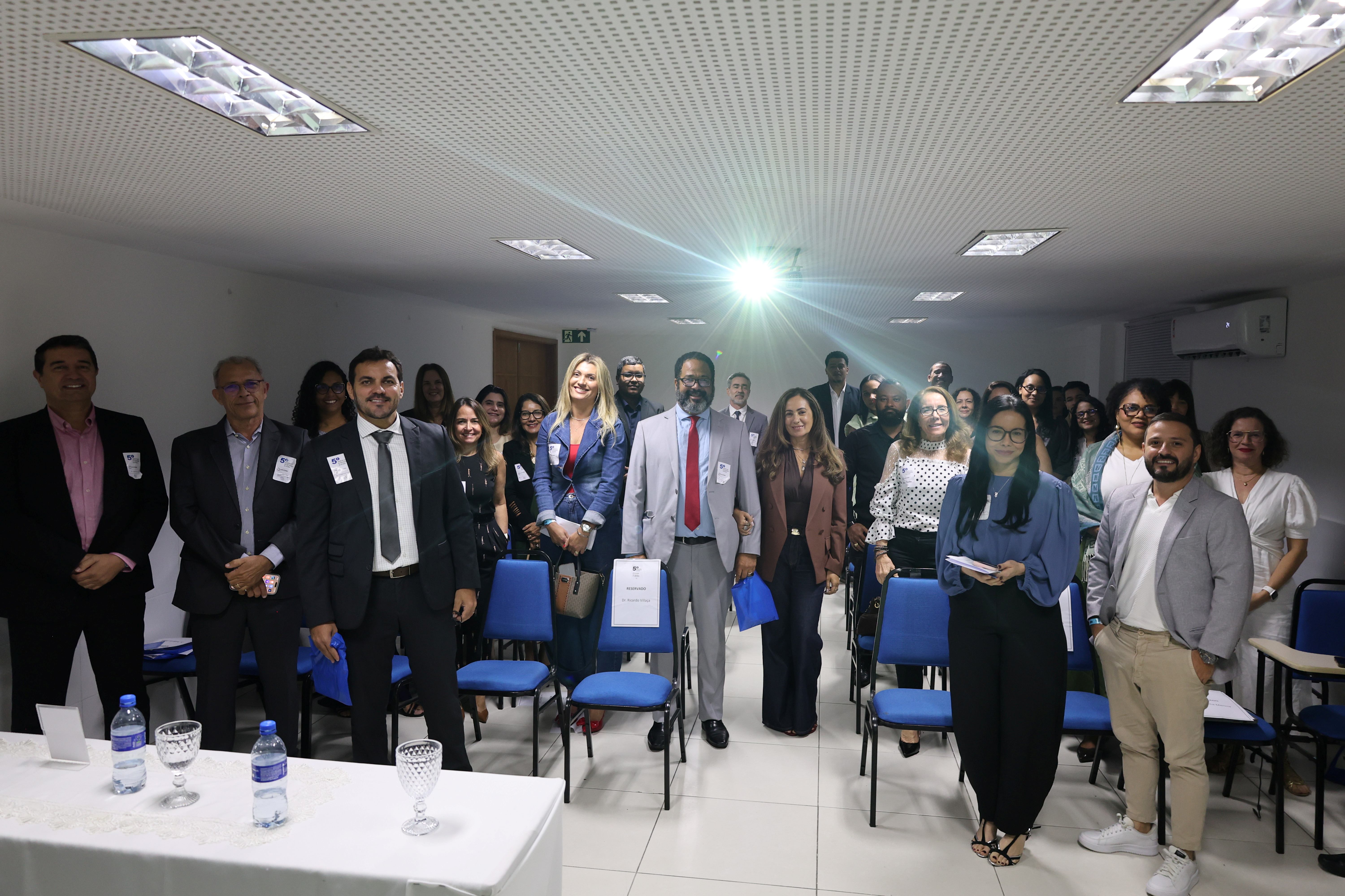 Fotografia de um grupo numeroso de pessoas reunidas em uma sala. Elas estão em pé e voltadas para a câmera, muitas sorrindo. A maioria usa roupas sociais, como ternos, blazers, camisas e vestidos, e algumas usam crachás. Ao centro, há um homem de terno cinza e gravata vermelha. À frente, há cadeiras azuis. No canto inferior esquerdo, uma mesa coberta com toalha branca exibe garrafas de água e taças. O ambiente tem teto com iluminação embutida e ar-condicionado.