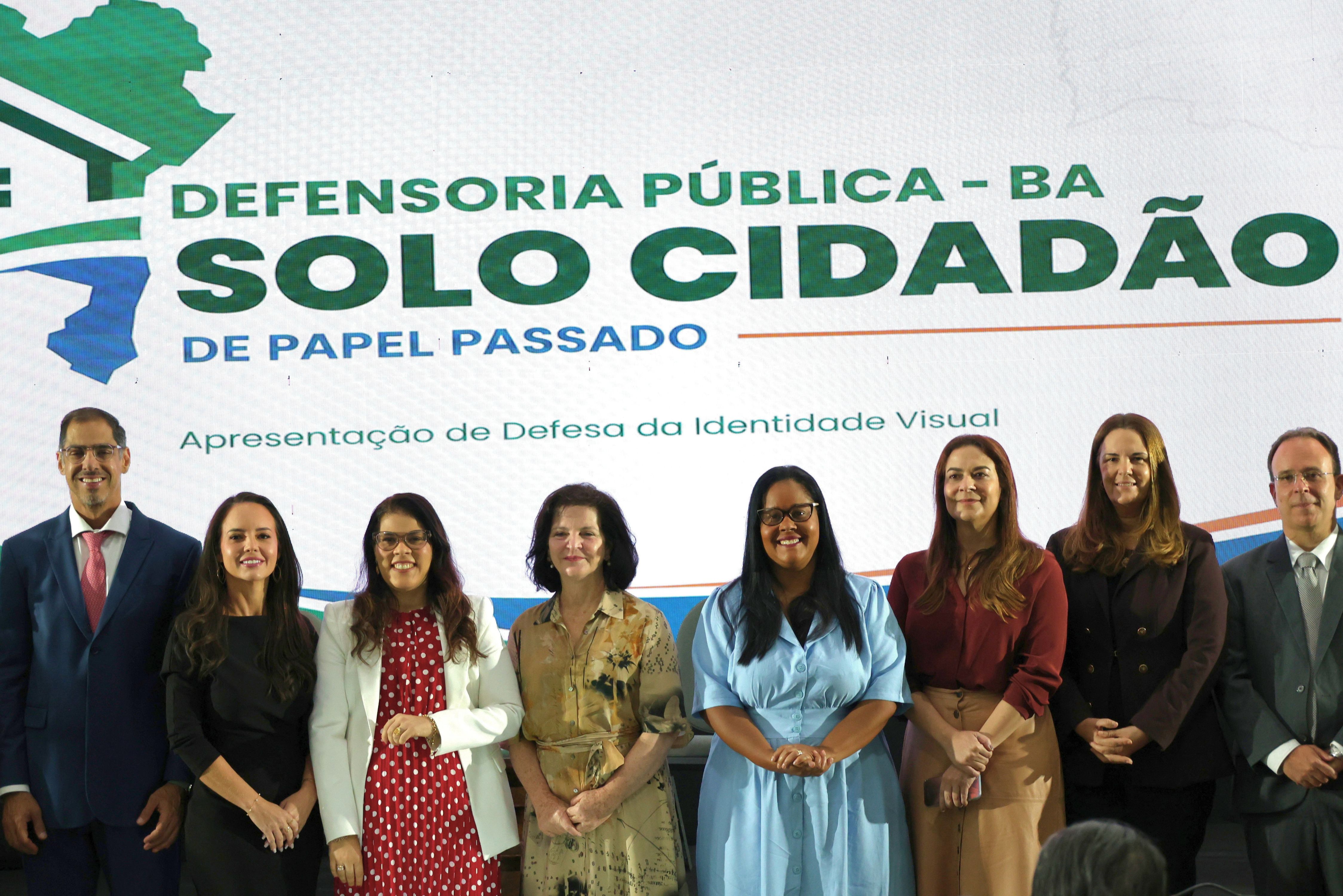 Em frente a um painel com o texto “Defensoria Pública – BA” e “Solo Cidadão – De Papel Passado”, sete pessoas posam para uma foto. Da esquerda para a direita, há três homens e quatro mulheres, vestidos formalmente. As mulheres usam roupas em preto, branco, vermelho, bege e azul-claro. Todos estão de pé, lado a lado, sorrindo ou olhando para a câmera. O painel também traz a frase “Apresentação de Defesa da Identidade Visual”.