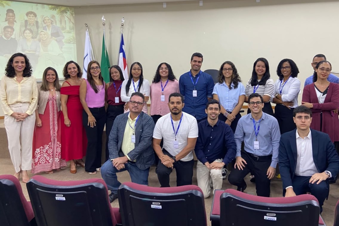  Fotografia de um grupo de cerca de vinte pessoas reunidas em uma sala de aula, posando para uma foto coletiva. Algumas estão em pé, formando duas fileiras ao fundo, e outras estão sentadas ou agachadas à frente. Vestem roupas variadas, em diferentes cores, e muitas usam crachás com cordões azuis. Ao fundo, há três bandeiras e uma projeção na parede com a imagem de outras pessoas. Na parte inferior, aparecem fileiras de cadeiras estofadas em tons de vinho.
