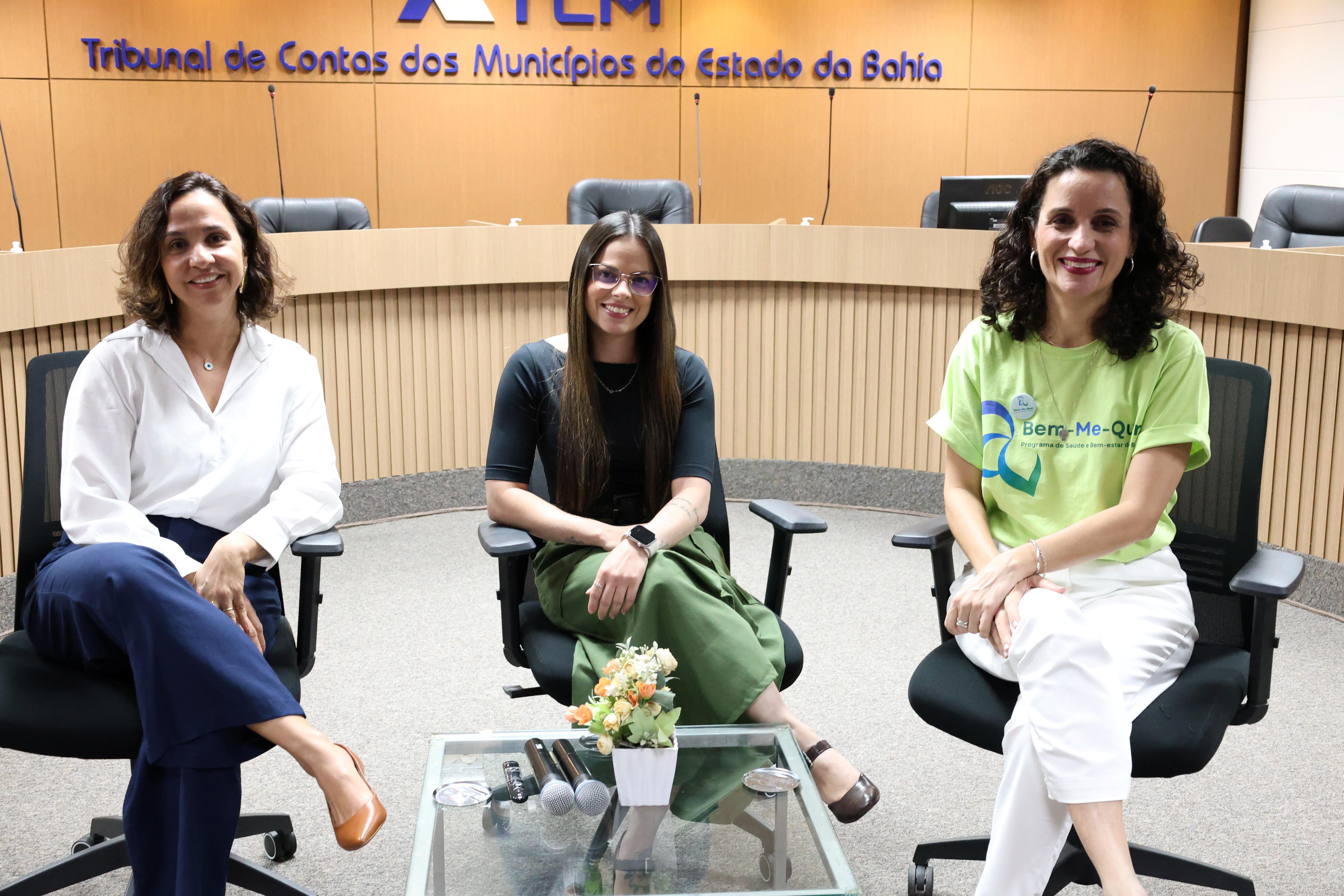 Fotografia de três mulheres sentadas em cadeiras, em um ambiente institucional. A mulher à esquerda usa camisa branca, calça azul-marinho e sapatos marrons. A mulher ao centro veste blusa preta, calça verde, óculos e relógio. A mulher à direita usa camiseta verde-clara com estampa, calça branca e tênis. Todas sorriem para a câmera. À frente delas, há uma mesa de vidro com microfones e um pequeno arranjo de flores. Ao fundo, vê-se um painel de madeira com a identificação do TCE-BA
