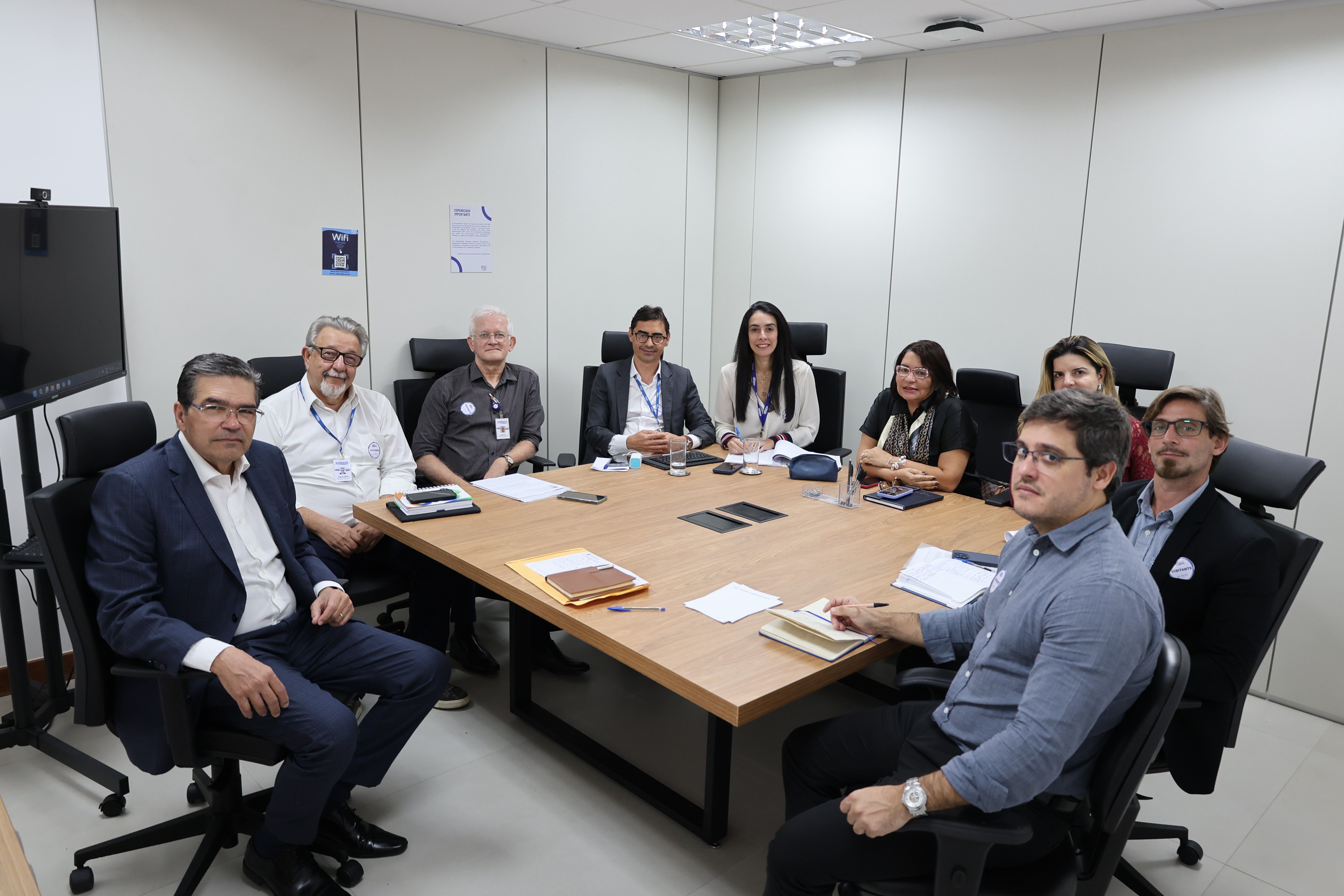 Sala de reunião com paredes claras, mesa retangular de madeira e cadeiras pretas. Nove pessoas estão sentadas ao redor da mesa, voltadas para a câmera. Algumas usam crachás e estão com papéis, cadernos, celulares e canetas sobre a mesa. Ao fundo, há uma tela de televisão, cartazes nas paredes e luminárias no teto.