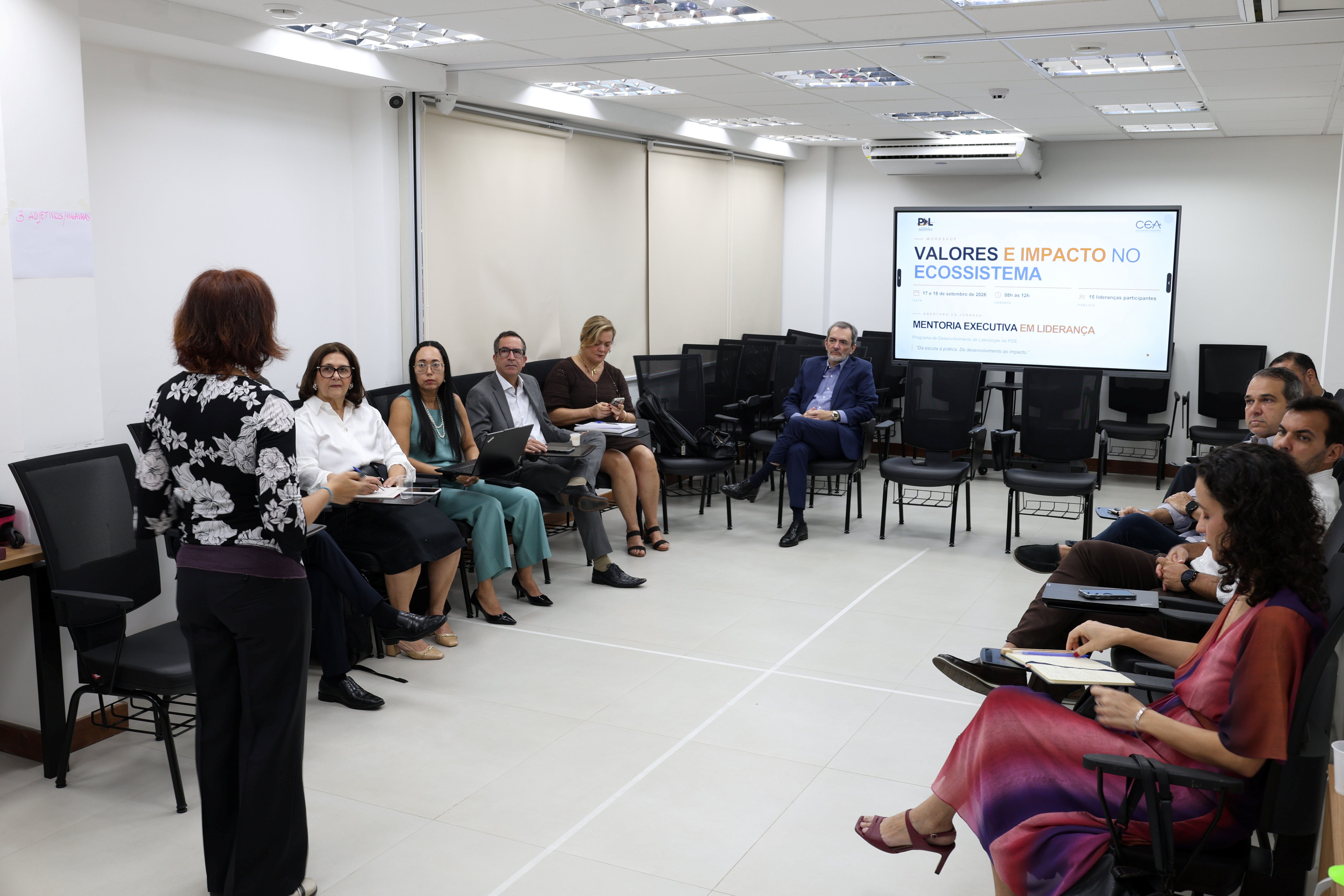 Fotografia de uma reunião em uma sala ampla e clara, com pessoas sentadas em cadeiras dispostas em semicírculo. Ao centro, uma mulher está em pé, de costas para a câmera, conduzindo a atividade. Os participantes fazem anotações e utilizam computadores. Ao fundo, há uma tela exibindo a apresentação “Valores e Impacto no Ecossistema” e informações sobre mentoria executiva em liderança.
