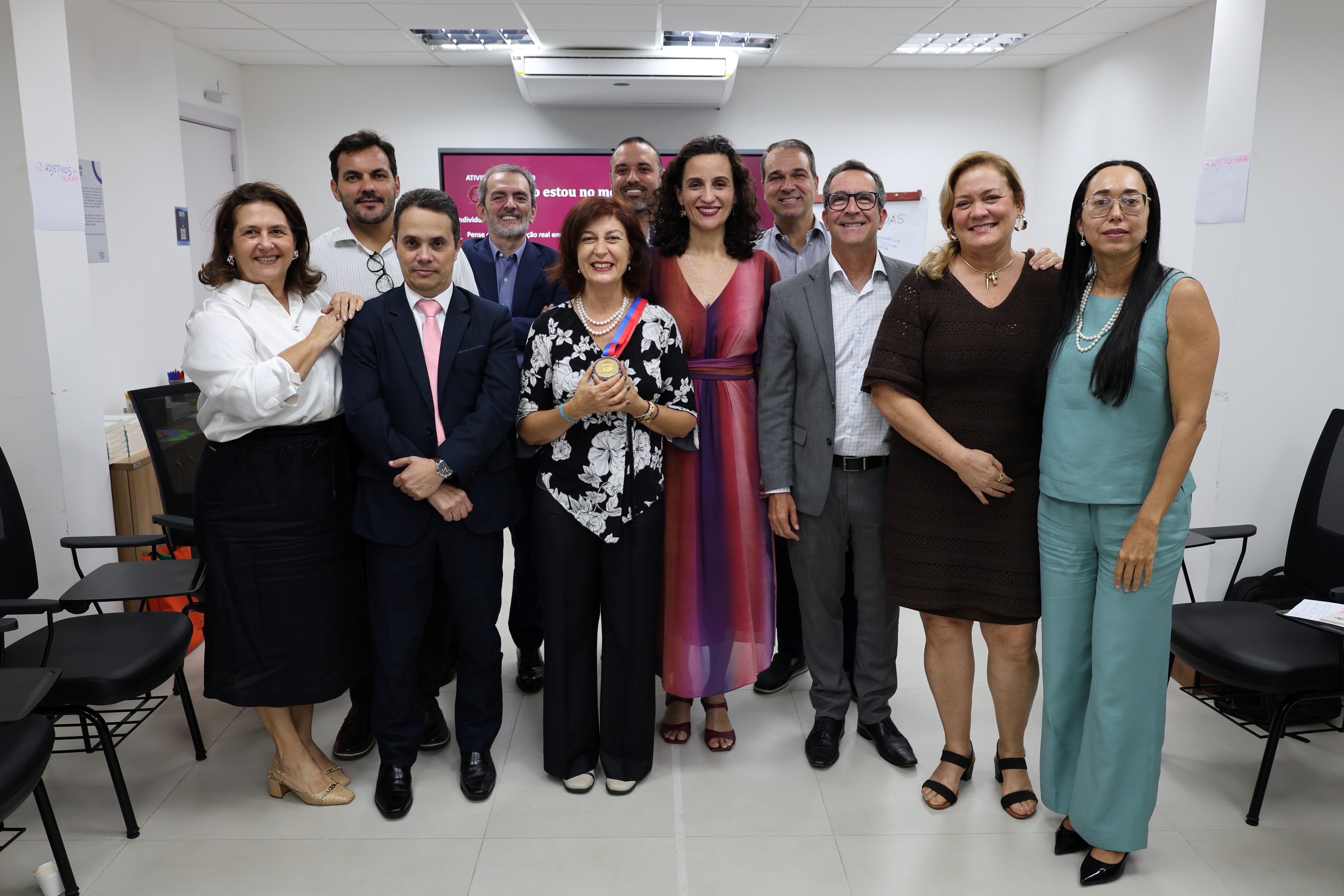 Fotografia de um grupo de onze pessoas reunidas em uma sala. Elas estão em pé, lado a lado, sorrindo e olhando para a câmera. Ao centro, uma mulher usa blusa preta com estampa de flores brancas, colar de pérolas e segura uma medalha. Os demais participantes usam roupas sociais e casuais. Ao fundo, há uma tela com uma apresentação e cadeiras organizadas pela sala.
