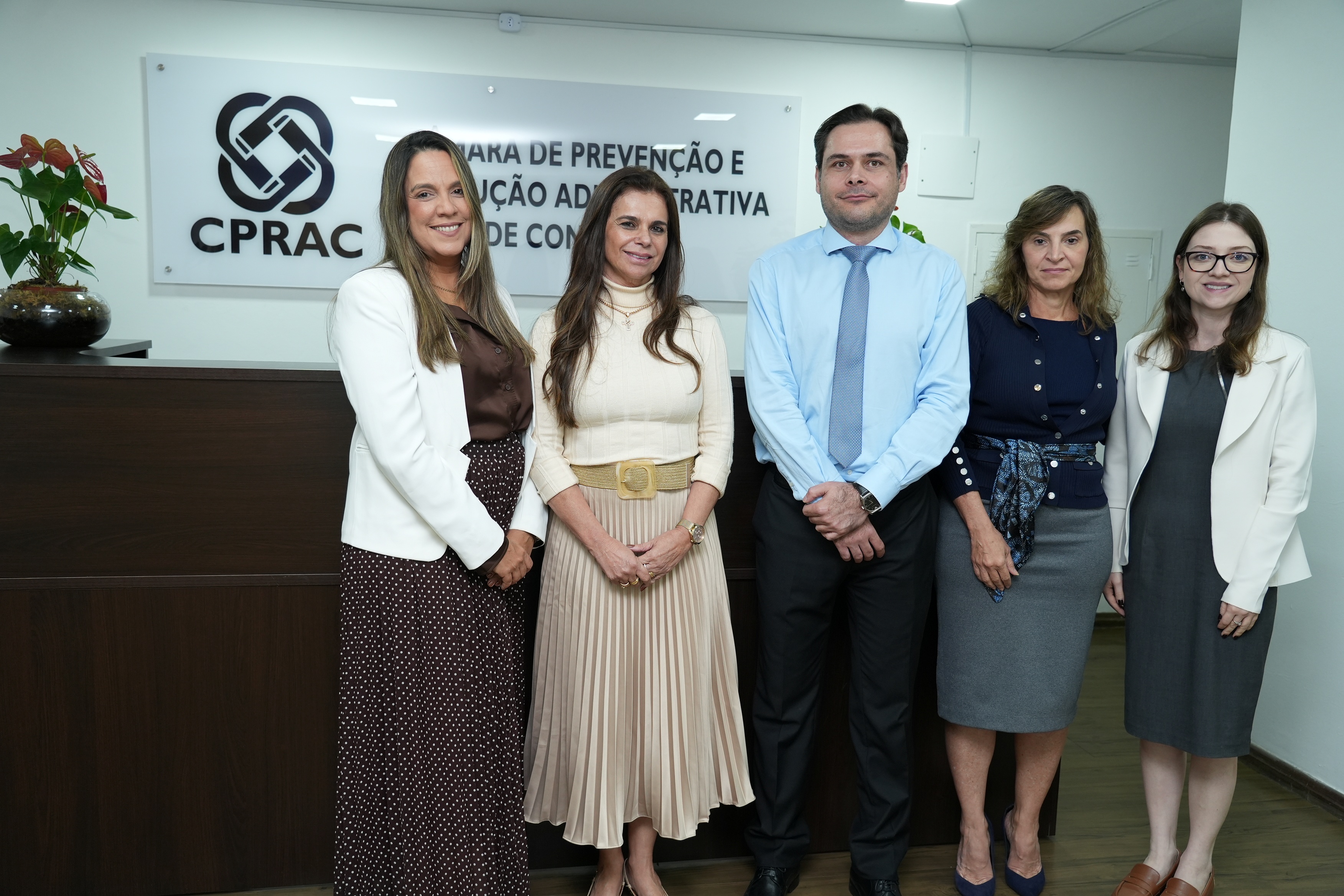 Foto de cinco pessoas em pé, lado a lado, em um ambiente institucional. Ao fundo, há um balcão de madeira e uma placa com o logotipo e a sigla “CPRAC”. Da esquerda para a direita, estão duas mulheres, um homem e duas mulheres. As mulheres usam roupas sociais, com blazers, saias e vestidos. O homem veste camisa azul-clara, gravata estampada e calça escura. Todos olham para a câmera e sorriem discretamente.