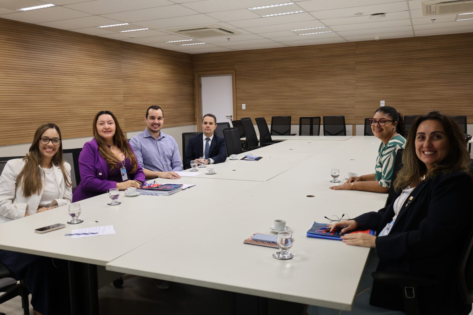 Sala de reuniões ampla, com paredes revestidas de madeira e uma grande mesa branca. Seis pessoas estão sentadas ao redor da mesa, em lados opostos, voltadas para a câmera. Há quatro mulheres e dois homens, todos vestidos de maneira formal ou social. Sobre a mesa, há copos com água, xícaras, celulares, cadernos e documentos. Ao fundo, há cadeiras pretas e uma porta.