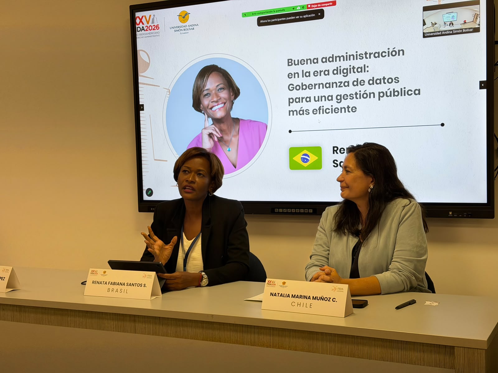 Fotografia de um painel de evento. Duas mulheres estão sentadas à mesa. À esquerda, uma mulher negra, de cabelos curtos, usa blazer preto e blusa clara. À direita, uma mulher de cabelos longos usa blusa cinza. Ao fundo, um telão exibe o título “Boa administração na era digital: Governança de dados para uma gestão pública mais eficiente”, além das imagens e nomes das participantes. Sobre a mesa, há placas de identificação, microfones e objetos pessoais.