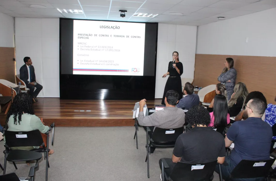 Uma palestrante de vestido preto fala ao microfone no palco, apontando para um telão que detalha informações sobre "LEGISLAÇÃO" e "PRESTAÇÃO DE CONTAS". Dois outros palestrantes acompanham a fala nas laterais do palco.