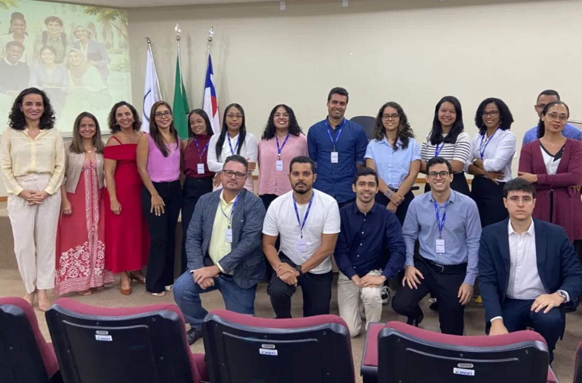  Fotografia de um grupo de cerca de vinte pessoas reunidas em uma sala de aula, posando para uma foto coletiva. Algumas estão em pé, formando duas fileiras ao fundo, e outras estão sentadas ou agachadas à frente. Vestem roupas variadas, em diferentes cores, e muitas usam crachás com cordões azuis. Ao fundo, há três bandeiras e uma projeção na parede com a imagem de outras pessoas. Na parte inferior, aparecem fileiras de cadeiras estofadas em tons de vinho.