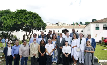Grupo de cerca de vinte pessoas posa para uma foto ao ar livre. Elas estão organizadas em duas fileiras, em pé, sobre um pequeno palco. A maioria veste roupas sociais, como ternos, camisas e vestidos. Ao fundo há um prédio histórico de dois andares, com paredes claras, janelas verdes e telhado de telhas claras. Bandeiras estão hasteadas no centro do edifício. À esquerda há uma grande árvore com copa cheia e, ao redor, um jardim com gramado. Algumas pessoas aparecem ao fundo caminhando pelo espaço.