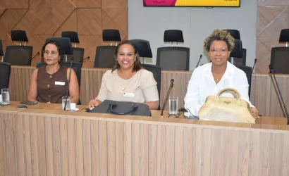 Três mulheres estão sentadas lado a lado em uma mesa de reunião, olhando para a câmera. Sobre a mesa há bolsas, copos de água e equipamentos de áudio. Ao fundo aparecem cadeiras e um painel de madeira na parede.