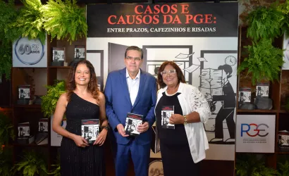Uma foto em grupo com três pessoas de pé e sorrindo, segurando exemplares de um livro. Da esquerda para a direita, temos uma mulher de vestido preto longo de um ombro só, um homem de terno azul escuro e uma mulher de blazer branco e blusa preta. O fundo é composto por um painel preto com texto e ilustrações em branco, prateleiras de madeira com livros e vasos de plantas, e o logotipo da PGE com a inscrição "PROCURADORIA GERAL DO ESTADO DA BAHIA" e "60 ANOS".
