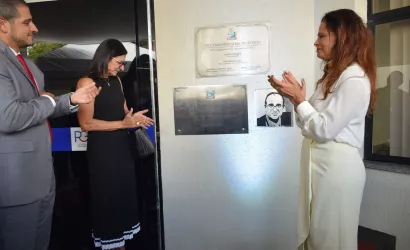 Três pessoas em um evento oficial. À esquerda, um homem de terno cinza gesticula; ao centro, uma mulher de vestido preto aplaude; à direita, uma mulher com camisa branca e lenço também aplaude. Eles estão diante de placas de inauguração na parede.