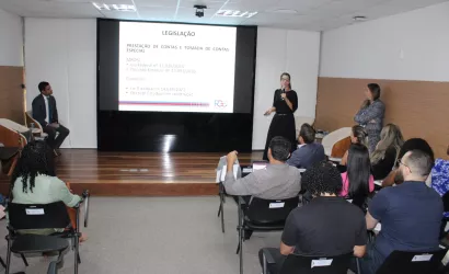 Uma palestrante de vestido preto fala ao microfone no palco, apontando para um telão que detalha informações sobre "LEGISLAÇÃO" e "PRESTAÇÃO DE CONTAS". Dois outros palestrantes acompanham a fala nas laterais do palco.