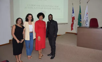 Quatro pessoas posam juntas para uma foto no palco do auditório, em frente à projeção do curso. Da esquerda para a direita: uma mulher de vestido preto, uma mulher de blazer claro e jeans, uma mulher de vestido vermelho e um homem negro de terno escuro.
