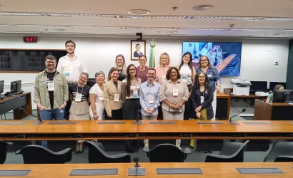 Foto de um grupo de cerca de 14 pessoas reunidas em uma sala de reuniões. Elas estão de pé, lado a lado, atrás de uma longa mesa de madeira com microfones. Ao fundo, há bandeiras, um retrato, um relógio digital e uma tela exibindo outra imagem da sala. Todos posam para a foto e usam crachás de identificação.