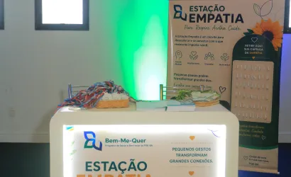 Foto de um espaço de divulgação chamado “Estação Empatia”. Ao centro, há um balcão iluminado, com cartazes sobre empatia, acolhimento, conexão e bem-estar. Sobre o balcão, estão cestas com materiais e pequenos cartões. Ao fundo, um painel vertical traz o mesmo tema, com uma flor, folhas e um suporte com cápsulas de papel. O ambiente é interno, com paredes claras e iluminação decorativa em verde e azul.