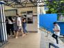 Ambiente de entrada de um prédio, com cobertura metálica azul e piso de concreto. À esquerda,  pessoas entram por um portão azul.  Ao centro, uma mulher de blusa preta e calça jeans caminha em direção ao portão. Atrás um homem de camisa polo branca, calça jeans e tênis sobe as escadas enquanto olha para um celular e segura um papel. Ao fundo, há paredes azuis, janelas, avisos fixados na parede e árvores visíveis acima do muro. O local é iluminado pela luz do dia.