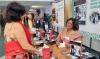 Foto de um estande de evento. À direita, uma mulher de óculos, usando roupa social em tom salmão e crachá, está sentada à mesa, sorrindo e autografando um livro. À esquerda, outra mulher, de costas para a câmera, usa roupa vermelha e carrega uma bolsa da mesma cor. Sobre a mesa, há vários exemplares do livro “Casos e causos da PGE-BA: entre prazos, cafezinhos e risadas”. Ao fundo, outras pessoas conversam e há painéis com materiais de divulgação.