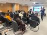 Sala de reunião com várias pessoas sentadas em cadeiras diante de uma tela. Ao fundo, um homem de terno segura um microfone e fala para o grupo. Outro homem está em pé próximo à tela, que exibe uma apresentação. As paredes têm revestimento de madeira, há janelas com persianas, ar-condicionado e projetor no teto. Os participantes observam a apresentação e alguns usam celulares ou computadores.