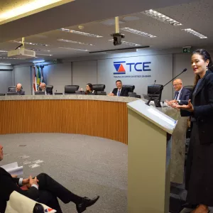Imagem mostra uma mulher em pé falando ao microfone, posicionada atrás de um púlpito no plenário do Tribunal de Contas do Estado da Bahia (TCE-BA). Ao fundo, sete pessoas estão sentadas em uma bancada semicircular, com laptops e microfones à frente. Todos estão com trajes formais. Três homens sentados na plateia observam a oradora com atenção. Na parede, há o logotipo do TCE e bandeiras ao fundo. O ambiente é bem iluminado, moderno e com clima institucional.