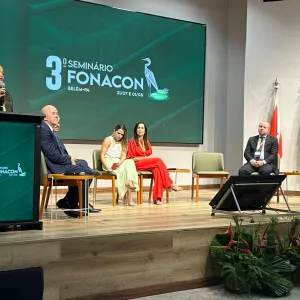 Mulher fala ao microfone em pé, atrás de um púlpito, no palco de um auditório. Ao fundo, uma grande tela exibe “3º Seminário FONACON – Belém-PA – 31/07 e 01/08”. Sentados ao lado da palestrante, há quatro pessoas: três mulheres e dois homens, todos atentos ao evento. À direita do palco, estão três bandeiras: do Pará, do Brasil e uma branca institucional. Na frente do palco, há plantas decorativas com folhagens verdes e flores vermelhas. O ambiente é iluminado, formal e organizado.