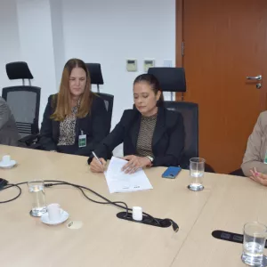 A imagem mostra um grupo de quatro pessoas em uma reunião ao redor de uma mesa. À esquerda, há um homem com cabelo grisalho, usando um terno cinza claro. Ao lado dele, uma mulher com cabelo longo e loiro, vestindo um blazer preto e blusa de estampa. No centro, uma mulher com cabelo preso, usando um terno preto, assina um documento. À direita, uma mulher de cabelo curto e grisalho, com óculos e acessórios coloridos, observa.