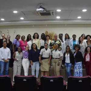 Um grupo diverso de pessoas posa sorrindo para uma foto em um auditório. Elas estão em pé sobre o palco e no chão à frente dele. A maioria são mulheres negras, duas usando uniformes, outras roupas sociais ou casuais. Ao fundo, há bandeiras e um arranjo decorativo com plantas secas. As cadeiras vinho do auditório aparecem na frente da imagem. A foto transmite um clima de encerramento de evento.