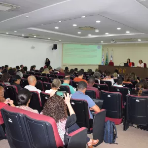 Auditório com várias fileiras de cadeiras ocupadas por pessoas assistindo a uma apresentação. Duas mulheres estão sentadas à mesa no palco, uma delas falando ao microfone. Atrás delas, há quatro bandeiras e uma projeção com texto na parede. O ambiente é iluminado com luzes no teto e tem aparência formal e organizada.