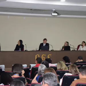 Em uma sala de auditório, várias pessoas estão sentadas assistindo a uma mesa de debate. Ao fundo, cinco pessoas estão sentadas atrás de uma bancada com microfones e laptops. No centro da mesa, um homem de terno fala ao microfone. As outras quatro pessoas, todas mulheres, ouvem atentamente. À esquerda da imagem, há uma bandeira do estado da Bahia. Na parte frontal da bancada está escrita a sigla "PGE". O ambiente é iluminado, e o público está atento à apresentação.