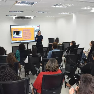 Palestrante apresenta conteúdo sobre Prevenção e Combate em uma sala com várias pessoas sentadas assistindo, olhando para uma tela grande ao fundo com slide colorido.