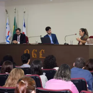 Em um auditório, três pessoas estão sentadas atrás de uma mesa participando de uma mesa-redonda. À esquerda, um homem de terno escuro e gravata vermelha está com as mãos entrelaçadas. No centro, outro homem de terno azul observa a mulher à direita, que fala ao microfone e gesticula com as mãos. Atrás deles, há três bandeiras (Brasil, Bahia e institucional). O público assiste, sentado em cadeiras acolchoadas.