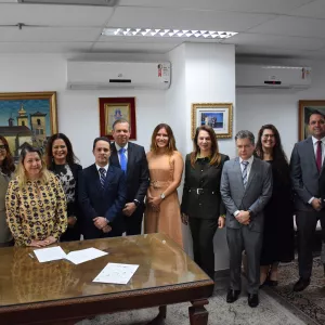 A imagem mostra um grupo de 12 pessoas em um ambiente interno. Eles estão posando para a foto, com 6 pessoas à frente e 6 atrás. As pessoas na frente estão em pé e algumas estão levemente curvadas para a frente, sorrindo. Ao fundo, há quadros nas paredes, e a mesa à frente delas tem documentos e objetos. Todos estão vestidos de forma formal.