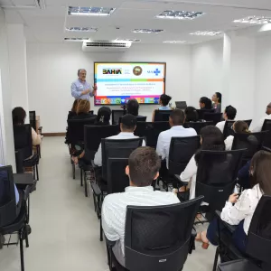 Em uma sala de reuniões clara e moderna, cerca de vinte pessoas estão sentadas, de frente para um palestrante que está em pé ao lado de uma tela. A apresentação exibe o tema “Assistência Farmacêutica no Estado da Bahia”, com logos do Governo da Bahia e do SUS. O público está atento, e a maioria veste roupas sociais. As cadeiras são pretas e a iluminação é branca, vinda do teto. A atmosfera é profissional e de aprendizado.