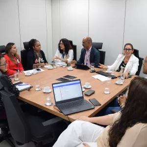 Imagem mostra dez pessoas reunidas em torno de uma mesa de madeira em uma sala de reunião. O grupo é formado por homens e mulheres, todos em trajes sociais. Algumas pessoas falam, enquanto outras escutam com atenção. Sobre a mesa, há copos com água, xícaras de café, celulares, cadernos, teclados e notebooks. Uma das telas do notebook está aberta em um sistema de e-mails. O ambiente é claro, com paredes brancas e cadeiras pretas, transmitindo um clima de reunião formal e colaborativa.