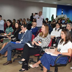 A imagem mostra uma plateia atenta em um auditório. As pessoas estão sentadas em cadeiras vermelhas com rodinhas, e a maioria usa roupas formais. Algumas estão anotando, outras assistindo e algumas tirando fotos com celulares. O ambiente é iluminado, com piso de madeira clara e uma grande imagem do Farol da Barra ao fundo. A cena transmite um clima de evento institucional ou reunião profissional.