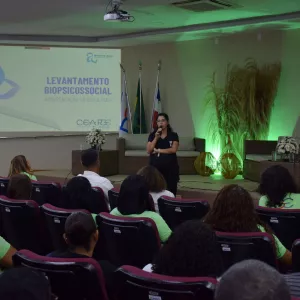 Auditório com público assistindo a uma mulher que está de pé no palco, falando ao microfone. Ela veste roupa preta e está posicionada entre sofás e arranjos decorativos com iluminação verde. Ao fundo, há três bandeiras e um telão exibindo o título da apresentação: “Levantamento Biopsicossocial – Apresentação do Resultado”.