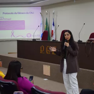 A imagem mostra uma mulher de cabelos longos falando ao microfone em auditório. Ela veste blazer marrom e calça branca. Ao fundo, no telão, aparece a apresentação intitulada “Protocolo de Gênero do CNJ”, com foto e informações da palestrante Daniela Portugal, doutora e mestre em Direito Público pela UFBA. Algumas pessoas sentadas nas poltronas vermelhas acompanham a palestra, enquanto a mesa principal tem bandeiras e microfones.