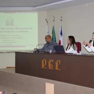 Na imagem, três pessoas estão sentadas atrás de uma mesa de auditório da PGE. À esquerda, um homem de camisa cinza olha para a frente. No centro, uma mulher de blazer branco sorri. À direita, outra mulher de blazer branco fala ao microfone com gestos expressivos. Atrás deles há quatro bandeiras. Na parede, um telão projeta o título da apresentação: “Dosimetria das sanções decorrentes de ilícitos em licitações e contratos”, com referência à lei estadual nº 14.634/2023 e ao decreto nº 23.113/2024.