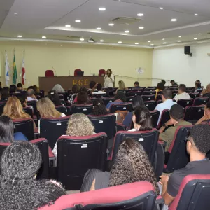 Visão mais ampla do auditório, com a plateia sentada em poltronas vermelhas. A palestrante aparece ao fundo, à frente da tela, explicando o conteúdo jurídico projetado. O local é bem iluminado e há grande participação do público.