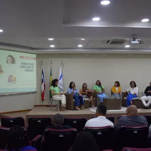 Foto de um auditório com um debate em andamento. No palco, sete pessoas estão sentadas em poltronas de vime, participando de uma roda de conversa. Uma das mulheres fala ao microfone. À esquerda, há um telão com o tema do evento: “Vamos falar sobre rede de cuidado?”. No fundo, há três bandeiras: da Bahia, do Brasil e uma branca institucional. O público está sentado em cadeiras vermelhas, assistindo à conversa. O ambiente é iluminado por luzes de teto.