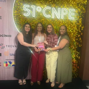 Na imagem, quatro mulheres estão sorrindo e posando juntas em frente a um painel iluminado com a inscrição “51º CNPE”. Elas estão de pé, uma ao lado da outra. A primeira mulher, à esquerda, usa um vestido preto e segura a mão de uma colega. A segunda, de cabelo longo e blusa branca, segura um livro nas mãos. A terceira mulher usa uma blusa vermelha e calça bege. A quarta mulher, à direita, usa um vestido verde e está sorrindo. O fundo é dourado e decorado.