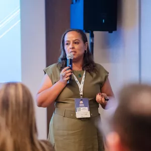 Close da palestrante, mulher de pele clara e cabelos castanhos soltos, usando vestido verde e crachá. Ela segura microfone e controle, enquanto fala ao público atento.