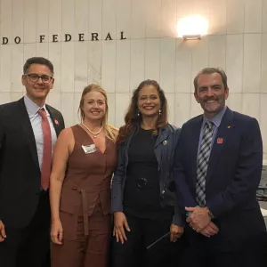 Quatro pessoas posam sorridentes em frente à parede com a inscrição “Senado Federal”. Estão bem-vestidas, demonstrando participação em um evento formal ou institucional. A imagem transmite profissionalismo, união e engajamento em atividades no ambiente legislativo.