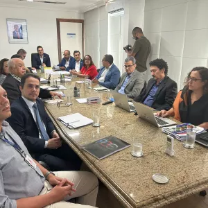 Reunião com 14 pessoas em uma sala de conferência. Homens e mulheres, em sua maioria de terno ou roupas sociais, estão sentados ao redor de uma mesa retangular. Dois notebooks estão abertos. Há copos com água, cadernos e documentos sobre a mesa. O ambiente é iluminado e com paredes brancas.