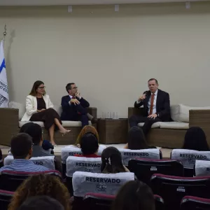 Três pessoas estão sentadas em poltronas no palco, participando de uma conversa. Uma mulher veste blazer claro e vestido vinho. Ao lado dela, um homem de terno azul e gravata vermelha. Outro homem, de terno escuro e gravata laranja, segura um microfone e fala. Atrás deles, há quatro bandeiras: da Bahia, do Brasil, da PGE-BA e uma quarta não identificada. À frente, o público assiste atento.