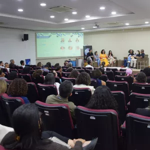 A imagem mostra um auditório com várias pessoas sentadas em cadeiras pretas de encosto alto, voltadas para a frente. No palco, à direita, há um grupo de oito pessoas sentadas em poltronas, participando de uma roda de conversa. Uma tela de projeção ao fundo exibe informações do evento, com fotos e nomes de participantes. À esquerda, o público observa atentamente. À direita, há um púlpito transparente e uma pessoa em pé, interpretando em Libras. O ambiente é iluminado por luzes de teto embutidas.