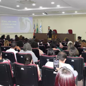 Auditório cheio de pessoas sentadas em poltronas vermelhas. À frente, um homem de blazer preto fala com microfone, enquanto no telão aparece a frase: “Afinal, não quero que ache um tédio a palestra”. Ao fundo estão as bandeiras do Brasil, da Bahia e outra institucional.