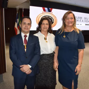 Três pessoas posam para foto em frente ao mesmo telão da homenagem. Um homem de terno azul, uma mulher de blusa branca e saia escura, e outra de vestido azul. Todos usam medalhas douradas e sorriem para a câmera.