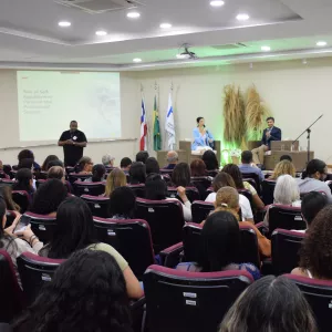 Auditório cheio, com pessoas sentadas em poltronas vinho, voltadas para o palco. No palco, duas pessoas estão sentadas em cadeiras de vime, conversando com microfones, enquanto um homem de preto faz interpretação em Libras no canto esquerdo. Ao fundo, há uma tela com apresentação escrita “Role of Self-Regulation in Personal and Professional Success” e bandeiras da Bahia, do Brasil e de uma instituição. Há também decoração com arranjos de capim seco e iluminação verde suave.
